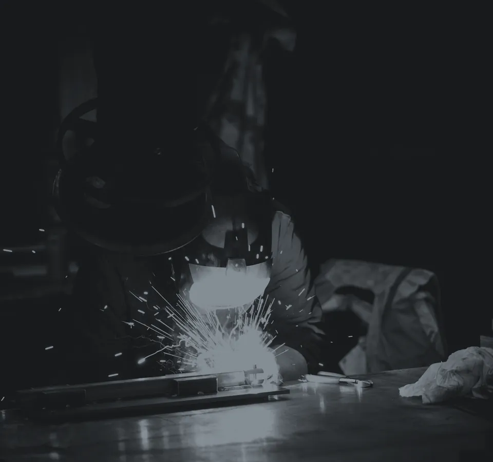 Welding in a metalworking shop: operator in a mask with sparks arcing across the bench.